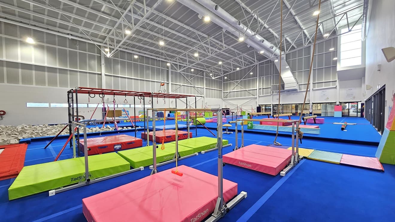 The floor is made of blue bouncy foam. Various gymnastics equipment such as balance beams, single bar trainers, horizontal bars, vault springboards, mats, and pommel horses are available. Brightly colored trampolines and tumbling blocks are also present. A foam pit with grey foams is positioned near the wall on the left. The high ceiling features an industrial look with exposed white metal beams.