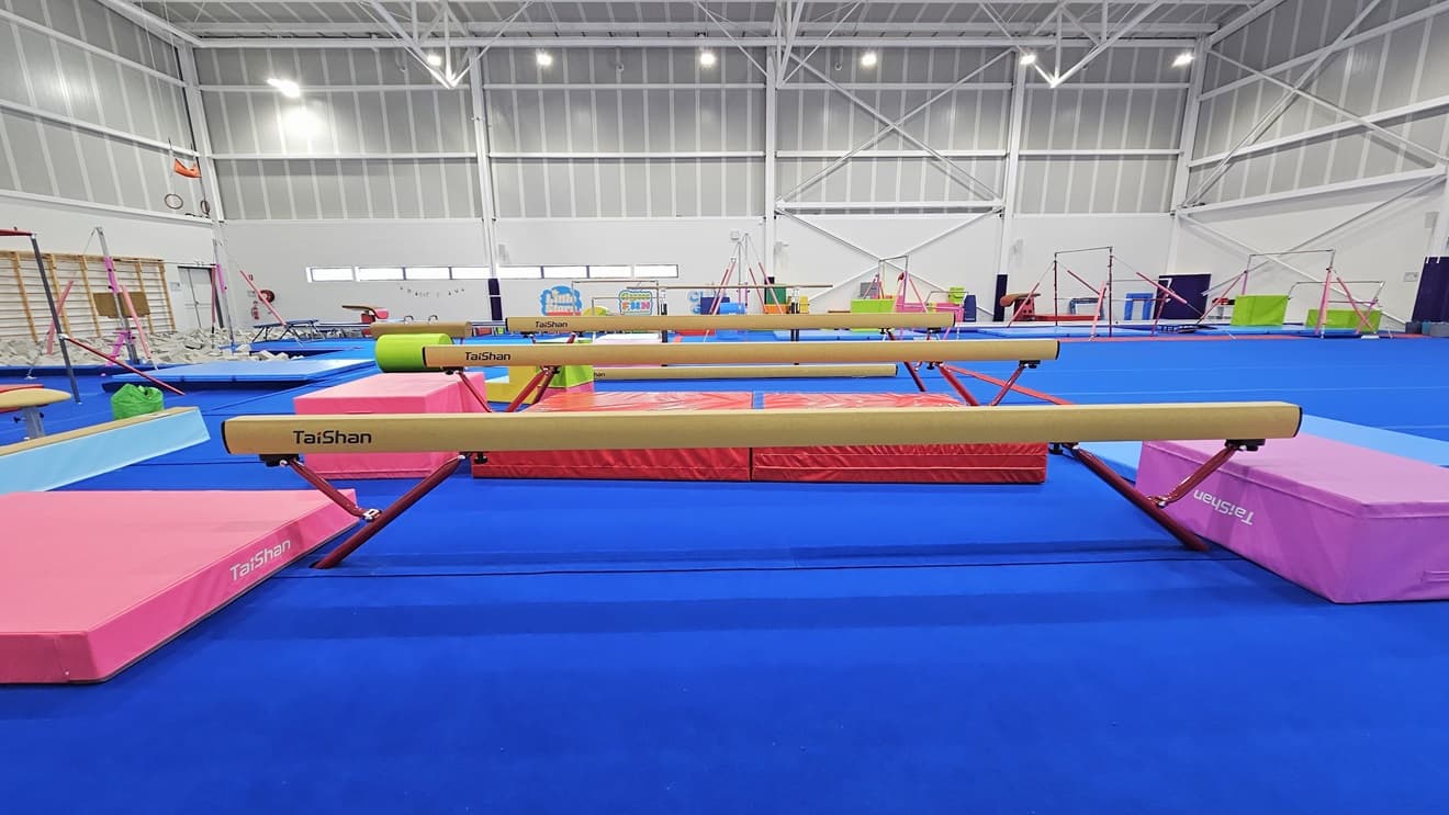 The floor is made of blue bouncy foam. Various gymnastics equipment, such as balance beams, is positioned in the middle. Other equipment like vault springboards and mats are available. Brightly colored trampolines and tumbling blocks are also present. A foam pit with grey foams is positioned near the wall on the left. The high ceiling features an industrial look with exposed white metal beams.