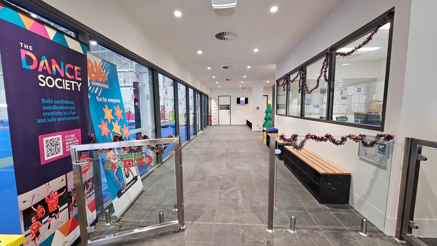 A controlled glass gate is located to the left of the reception. Huge retractable banners are positioned on the left. Inside, the corridor leads to the accessible bathrooms. To the left is the gymnastics area's glass wall, and to the right is a reception office. Christmas garland adorns the right window. There are two rubbish bins in place and a Christmas tree decoration. Further in is a door that leads to the Gymnastics area, and a flat-screen TV.