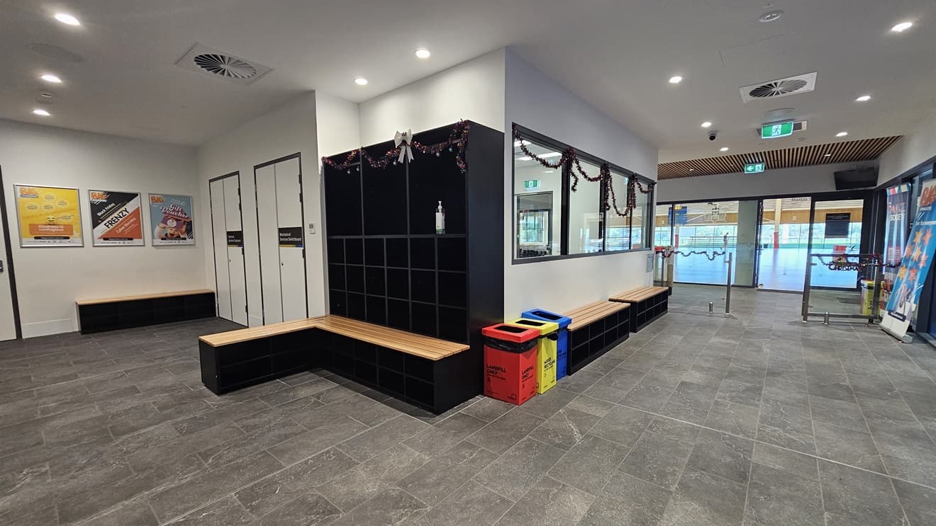 To the right is a controlled glass gate, and beside it are huge retractable banners. In the middle are bench seatings positioned against the wall. Rubbish bins are in place, along with a black storage rack for belongings adorned with Christmas garland. Two change rooms are available. The left wall features large poster banners. The floor has grey tiles, and the ceiling and wall are all white with LED lights.