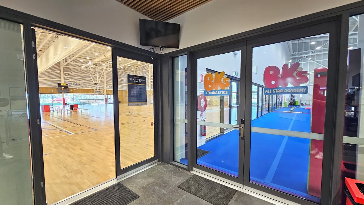 The entrance to the Sports Stadium is through a manual inward-opening glass sliding door. A poster is affixed to the wall. Non-slippery mats are placed on the flooring. To the right is a glass door entrance to BK's Gymnastics, equipped with stickers. A flat-screen TV is mounted in the upper corner. Inside the Stadium, the timber floor and the industrial-designed ceiling are visible.