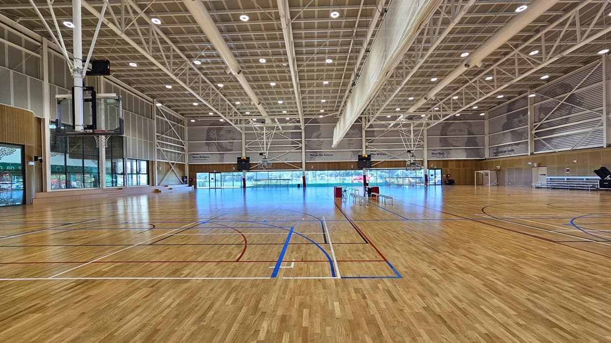 Inside are two basketball courts with timber floors, marked for various ball games. The stadium features timber floors and an industrial ceiling with exposed white beams, LED lights, and air conditioning vents. A screen net is positioned on the ceiling, serving as a separator between the two courts. Tables and chairs are arranged in the middle of the two courts. Distinctive paintings of sports legends adorn the upper wall. On the left is tiered timber seating, and on the right is tiered seating.