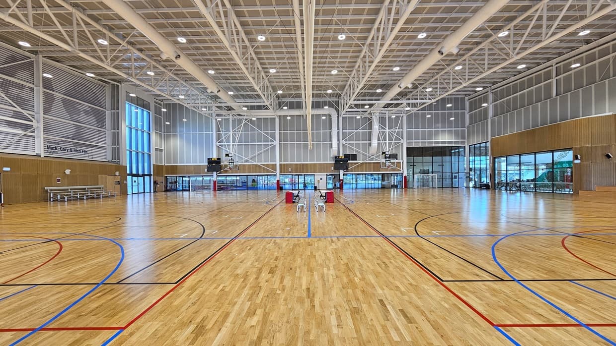 Inside, there are two basketball courts with timber floors, marked for various ball games. The stadium features timber floors and an industrial ceiling with exposed white beams, LED lights, and air conditioning vents. A screen net is positioned on the ceiling, serving as a separator between the two courts. Tables and chairs are arranged in the middle of the two courts. Distinctive paintings of sports legends adorn the upper left wall. Tiered seating is visible.