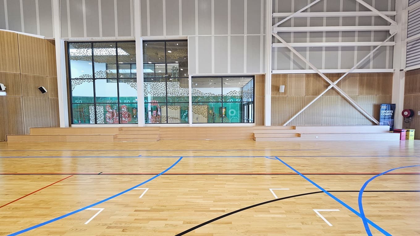 The stadium features timber floors marked for various ball games. The left and right walls have timber designs with mounted audio speakers. Rubbish bins and a fire hose reel, along with a retractable banner, are placed in the right corner. The middle glass features a foliage-shaped design in gold. Below it is a tiered timber seating.