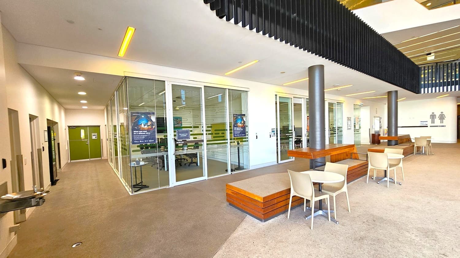 A modern indoor space with a beige carpeted floor. On the left, a corridor with green double doors and a water fountain. The middle features glass walls with posters, revealing a room with tables and chairs. On the right, there are round tables with beige chairs and wooden benches around black pillars. The ceiling has recessed lighting and black slatted panels. Restroom signs are visible in the background.