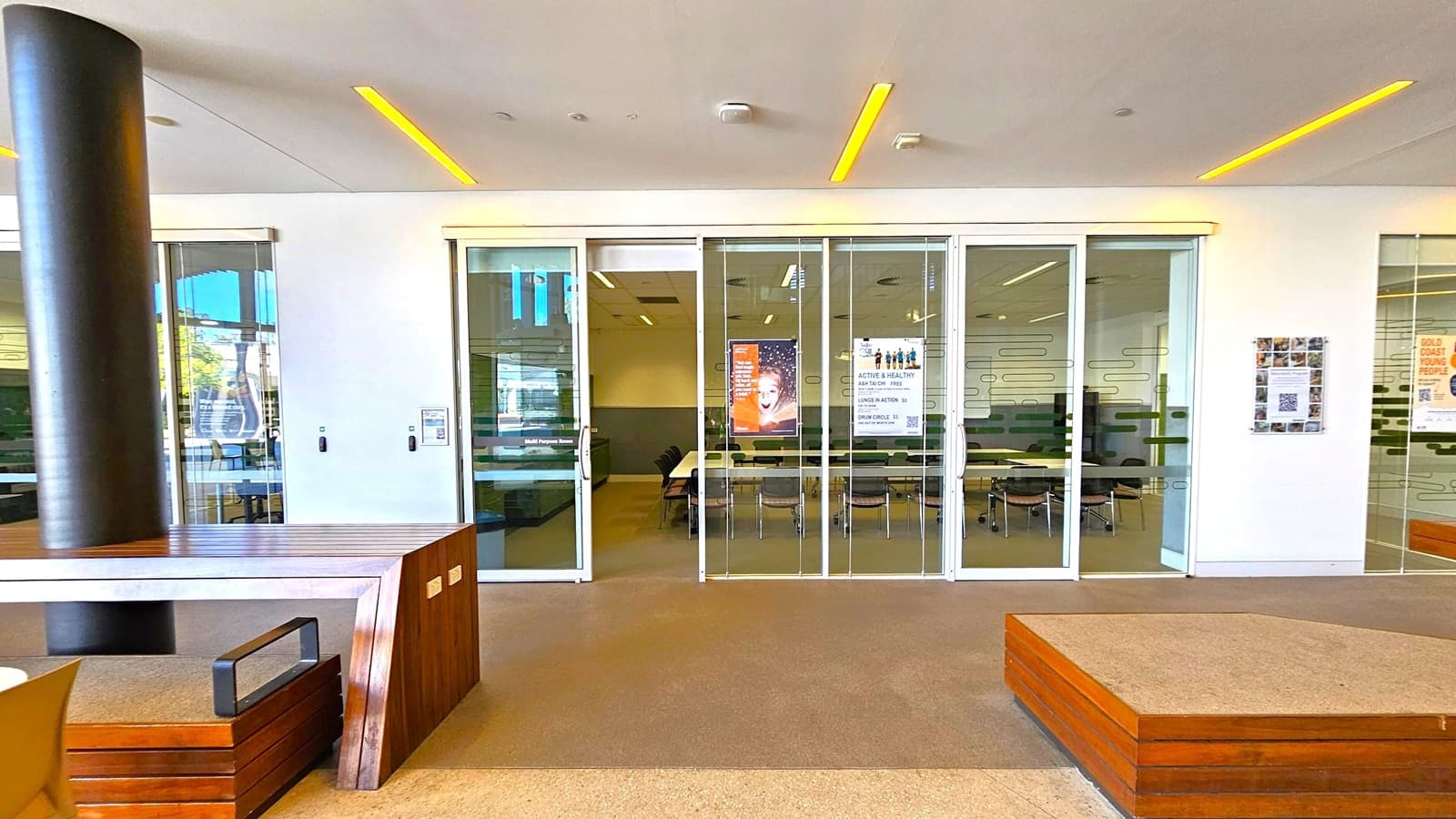 A modern interior space with a glass-walled room in the centre, featuring sliding doors. Inside, there are tables and chairs arranged in a meeting setup. Posters are visible on the glass, with one showing a smiling child. On the left, a dark cylindrical column and a wooden bench with a metal handle. On the right, a wall-mounted display with a QR code. The ceiling has recessed lighting, and the floor is carpeted in a neutral tone.