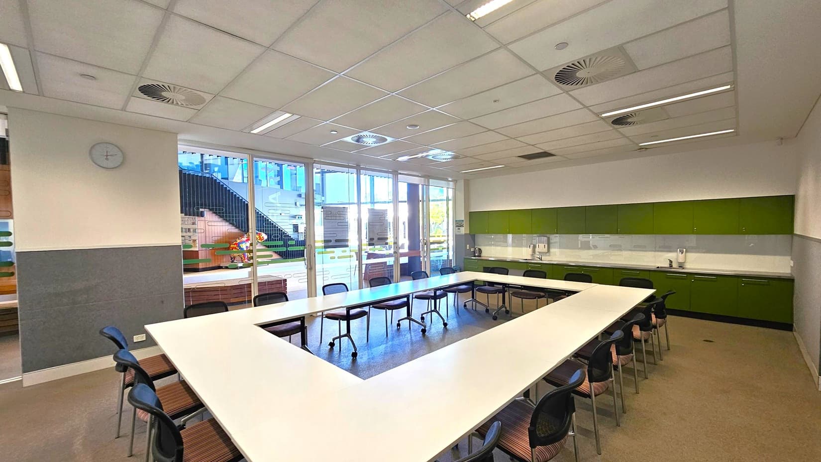 A room with a U-shaped arrangement of white tables and black chairs on a grey carpeted floor. The ceiling is fitted with white panels and circular vents. On the right, there is a long counter with green cabinets and a sink. The back wall features large glass doors with a view of an outdoor area, including stairs and colourful decorations. A clock is mounted on the left wall.