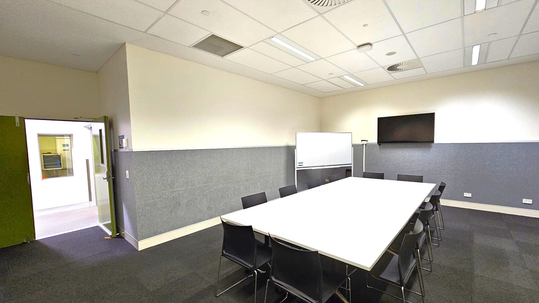 A conference room with a large rectangular table in the centre surrounded by black chairs. On the right wall, a flat-screen TV is mounted above a whiteboard. The walls are half grey and half cream, with a door open on the left leading to another room. The ceiling is fitted with fluorescent lights and ventilation panels. The floor is carpeted in dark grey tiles.