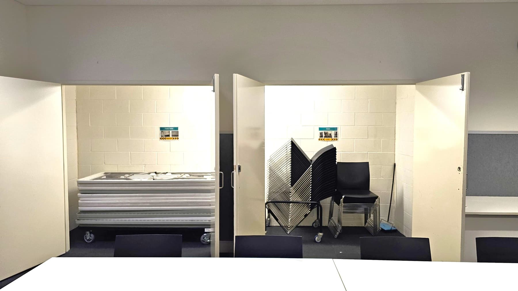 Two open storage areas with white walls. On the left, a stack of tables on wheels. On the right, a stack of chairs and folded metal racks. Both areas have small signs on the walls. The floor is dark, and the ceiling is light grey. In the foreground, a white table surface is partially visible.