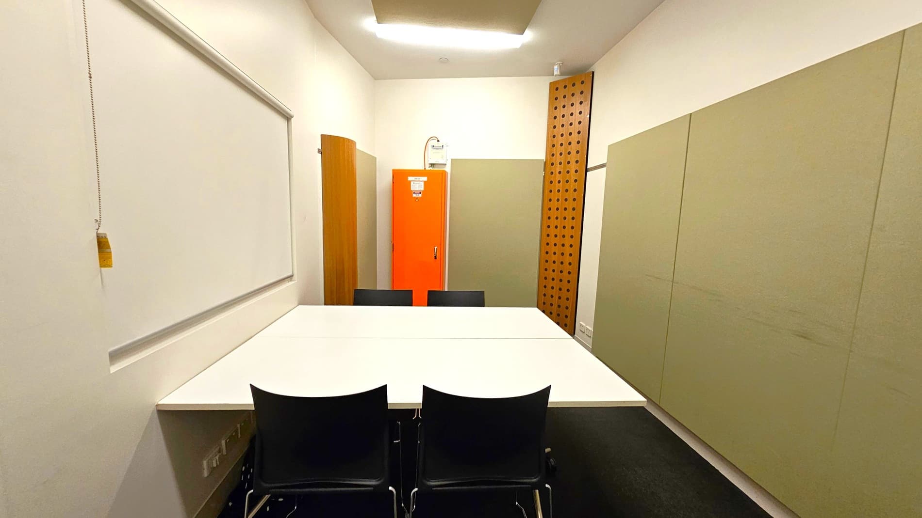 A small meeting room with a white table in the centre surrounded by four black chairs. On the left wall, there is a whiteboard with a pull chain. The back wall features an orange cabinet, flanked by beige panels and a wooden panel with circular cutouts. The floor is dark, and the ceiling has a rectangular light fixture.