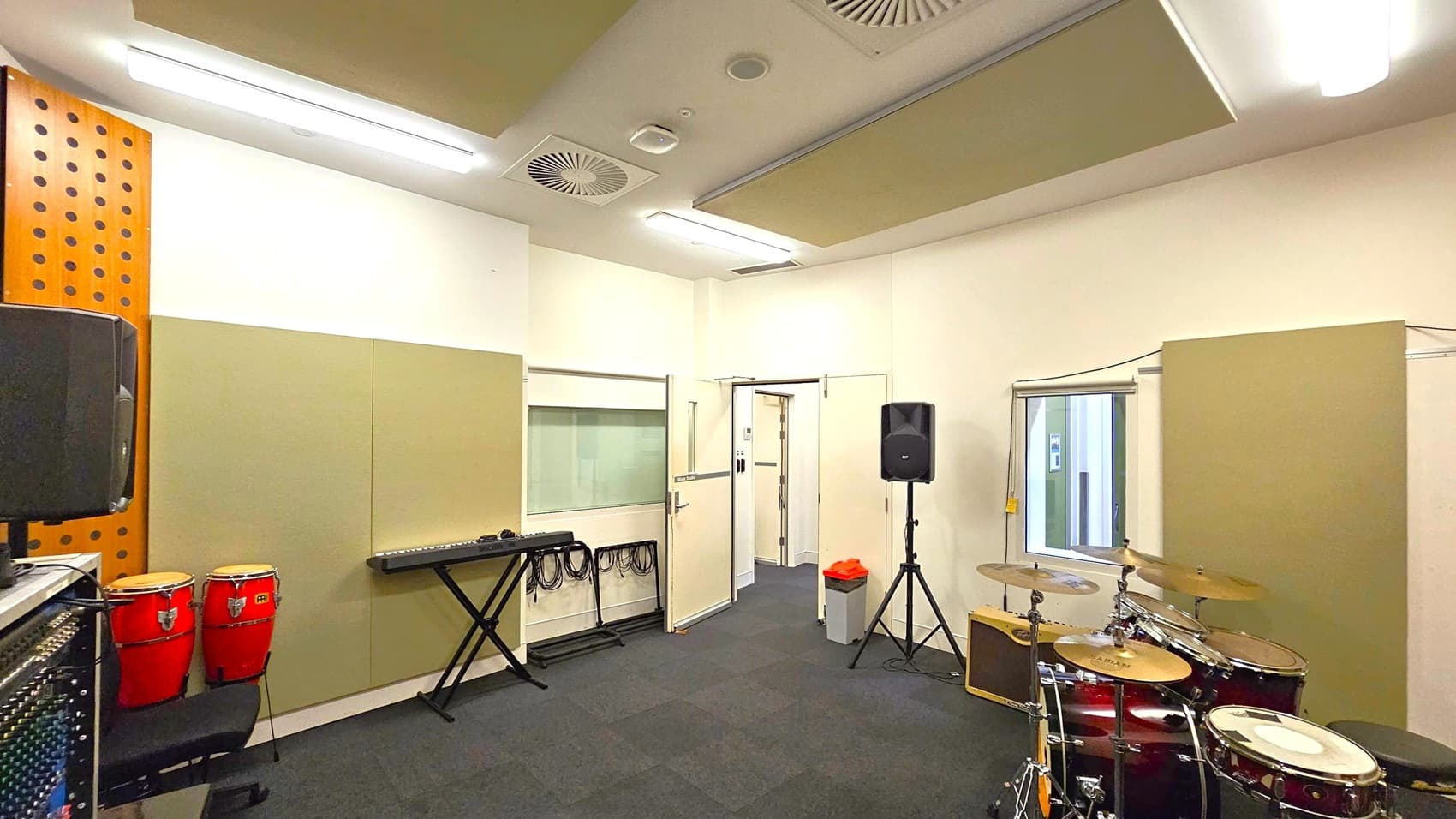 A music rehearsal room with a drum set on the right and a keyboard on a stand in the centre. On the left, there are red conga drums and a speaker. The walls are lined with beige acoustic panels, and there is a window with a blind partially open. The ceiling has fluorescent lights and ventilation panels. A door is open, leading to another room, and a waste bin is nearby. The floor is covered with dark carpet tiles.