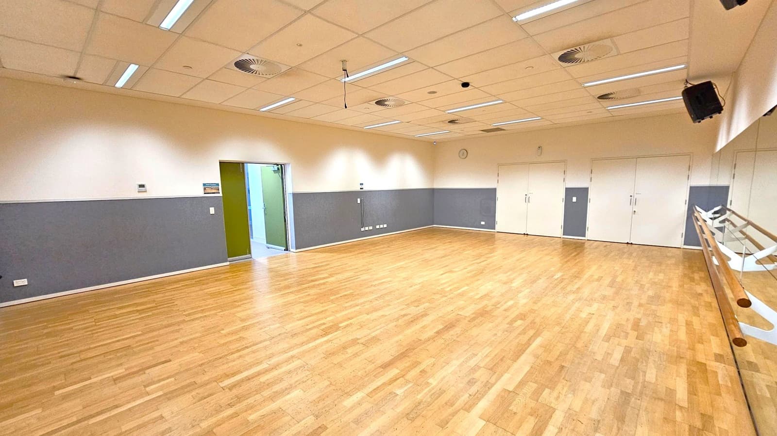 A spacious room with a wooden floor and a ceiling with recessed lighting and ventilation ducts. On the right, a mirrored wall with wooden ballet barres. The left wall is grey with a doorway leading to another area. The back wall has several white doors and a clock. The room is well-lit and appears to be a dance or exercise studio.