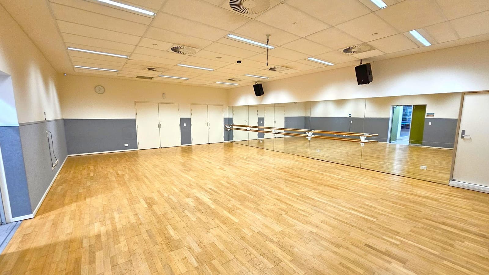 A spacious dance studio with a wooden floor and a mirrored wall on the right, featuring a ballet barre. The left wall is painted grey and white, with a few electrical outlets visible. The back wall has several closed doors. The ceiling is fitted with fluorescent lights and air vents. A speaker is mounted on the right wall. An open doorway on the right leads to another area.