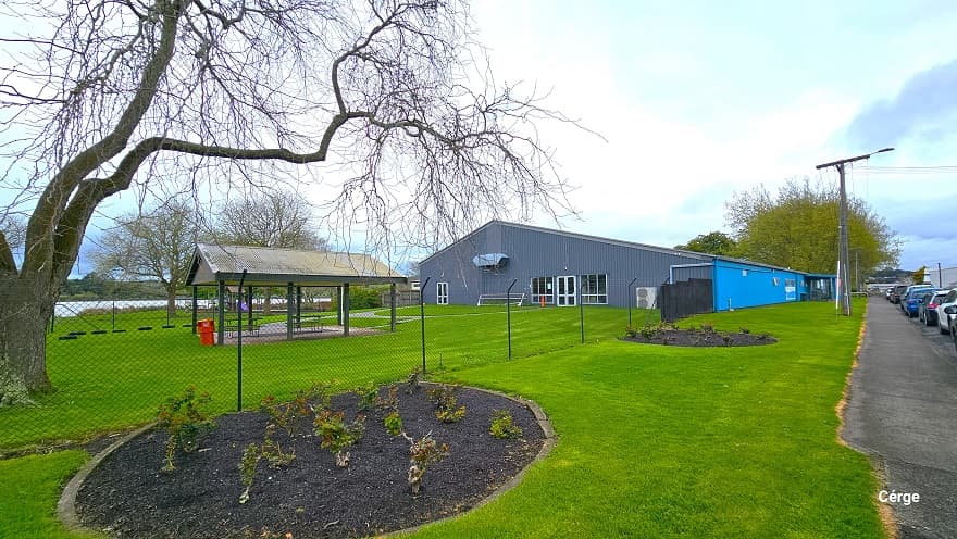 Surrounding the Huntly Aquatic Center is a spacious green field. The property is enclosed in a black, see-through, chain-link fence. Inside the property is a two-seating area with a roof, trees without much foliage, and a lake at the back. Right outside the fence are two circular gardens containing a variety of plants. A cemented pathway separates the parking space in front and the green field of the centre.