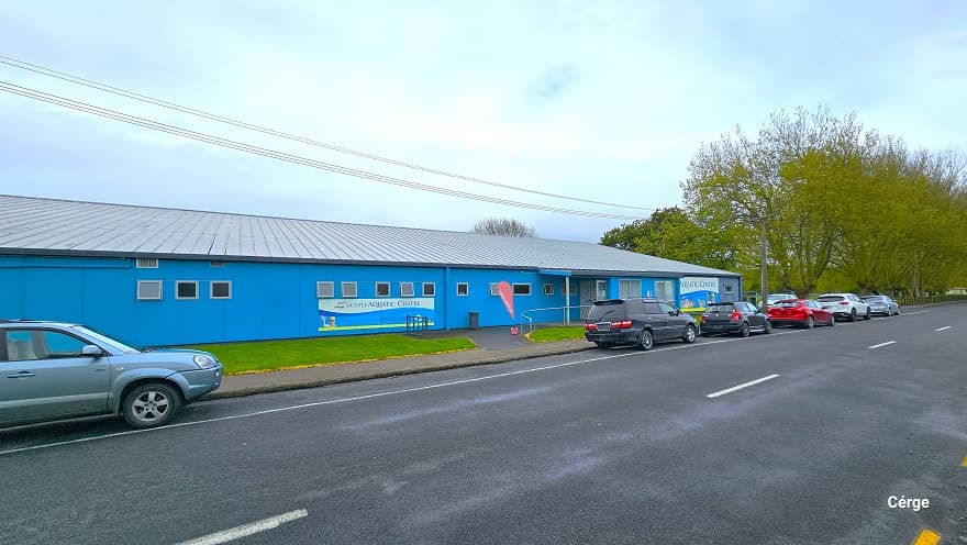 View of the parking area of the Aquatic Centre along the road of Wight Street, where several cars are parked. The low-set blue building can be seen in the background, with numerous trees located in the far right corner. There is a small white signage on the front blue wall detailing the name of the facility and a red, teardrop-shaped flag at the entryway to the facility.
