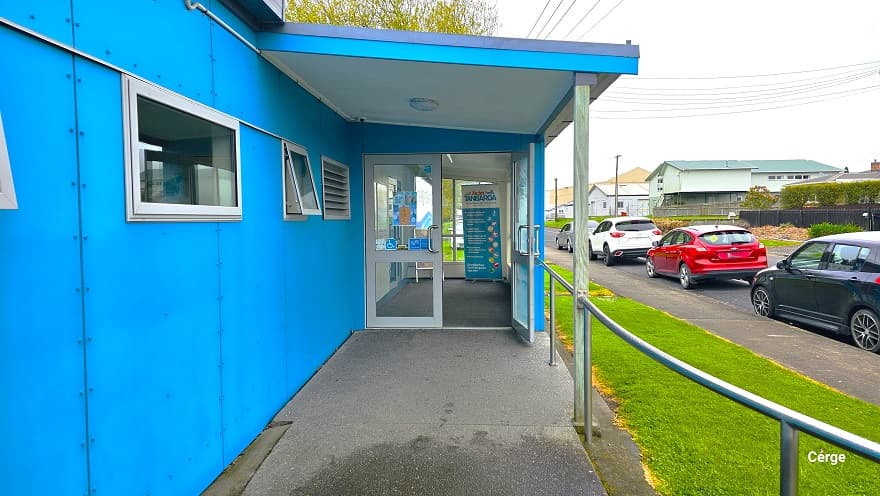 View of the entrance glass doors at Huntly Aquatic Centre coming from the cemented pathway with steel handrails. The main entrance has two glass doors lined with white painted panels, with the right door open. The small glass windows on the blue-painted wall to the left are slightly open. Grey, black, red, and white cars are parked in front. Residential buildings are noticeable in the far right corner of the image.