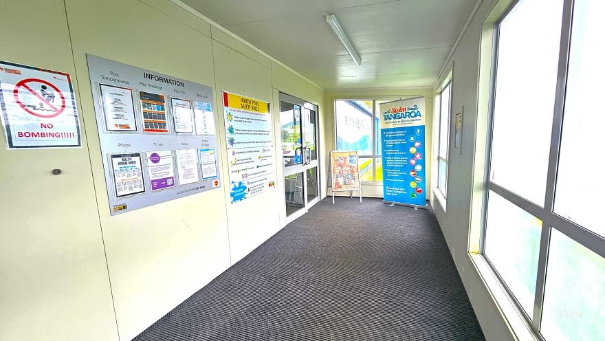 An inside look at the space after passing through the main entrance doors of Huntly Aquatic Centre. The floor is covered in a grey mat with a parallel line design. On the left wall are white, small signs for “No Bombing”, the information bulletin, and the Huntly Pool Safety Rules. On the right are tempered glass windows for privacy inside. Two advertising sign boards are placed in front of another entry door inside the centre.
