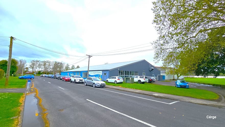 Opposite view of the entire front and side of the Huntly Aquatic Centre as well as the entire Wight Street. The front wall is painted in blue, while the right wall is painted in grey. The entire building has multiple glass windows. The grey wall has tall rectangular glass windows, while the front blue wall has a mixture of small and tall glass windows. Numerous vehicles are parked in front, each facing different directions. Power poles line both sides of the street.