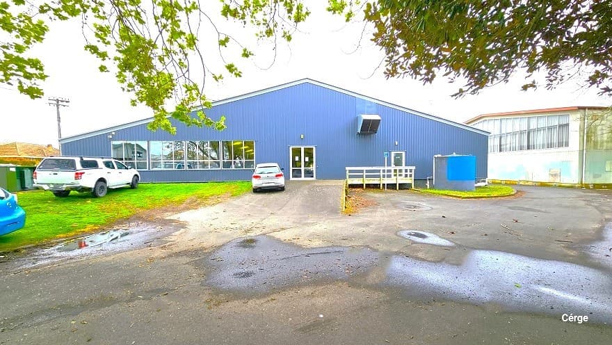 A closer view of the side angle of Huntly Aquatic Centre, which has a large cemented space and a landscaped area.. There are three cars outside, with two cars close to the building, while the blue car is near Wight Street. An inclined ramp leads to the glass entrance doors to the right is a small space with white-painted handrails. The glass windows reflect the natural surroundings of the centre. To the right is a large white building with glass windows near the roof.