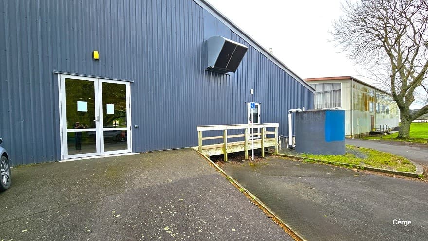 A closer look at the side entry glass doors leading inside the Huntly Aquatic Centre. The walls are painted in a dark purple hue. The inclined ramp leading to the glass doors is cemented. To the right is another glass door with white linings on the sides and white railings. There is a curvy elevation on the upper part of the wall for exhaust. Further to the right is a large circular tank with white PVC tubes, and to the back is a separate building.