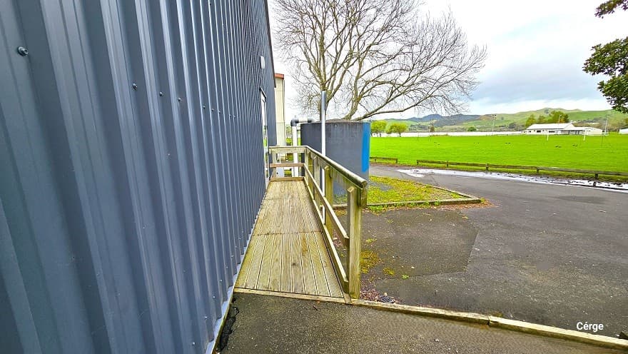 A closer look at the narrow space within the white railings of the side of Huntly Aquatic Centre. The floor space is made of wood as well as the handrails connected to the inclined cemented ramp. From this position, a large, open space of green field enclosed by small wooden fences can be seen. Towards the back is a huge tank with white PVC tubes, a large dead tree, and a glimpse of part of a white building to the back.