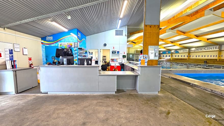 The image shows the double entrance glass doors, the reception desk, and the two pools. On the side of the reception desk are two young lifeguard males wearing orange and yellow uniforms. There is an adequate space that allows access surrounding each pool. Opposite to the two lifeguards is a pool with a 0.9-meter depth. The roof structure is made of corrugated iron and is well-lit with fluorescent lighting.