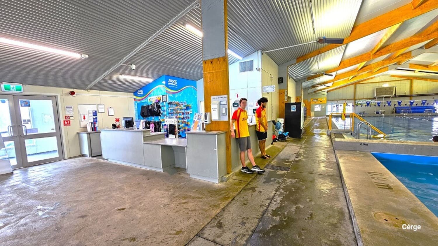 Front view of the white-to-grey reception desk area inside the Huntly Aquatic Centre. The reception area offers swimwear and other gear for sale. A water-like blue wall and a small office space at the back distinguish the reception area. On the left is a whiteboard with documents hanging next to a fire extinguisher. On the right are two pools with blue floating rope lanes, a string of blue triangular flags, and large, brown wooden frames that light the ceiling.
