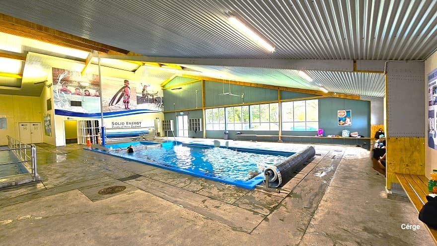 View of the small pool and children's splash area. Huge posters with kids swimming can be seen placed on top of the white wall to the back. A rolled waterproof sheet can be seen at one end of the pool. Near the exit glass doors are two hanging cabinets. Large glass windows on the side allow an appropriate amount of natural light to pass through and give natural scenery of the trees outside. Long brown benches are available throughout the surrounding area of the small pool.