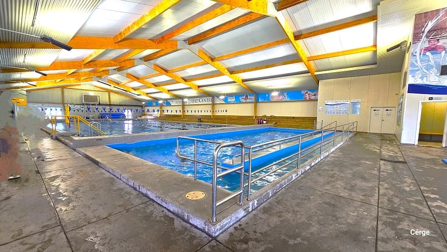A closer view of the ramp access with steel handrails of another small pool in Huntly Aquatic Centre. The ceiling has brown wooden support beams that support the white corrugated roof. Next to this small pool is a large pool with blue and white floating rope lanes. Four ladder accesses are marked by an inverted “U” steel frame on the pool sides. Long rows of low brown benches are placed near the large pool.
