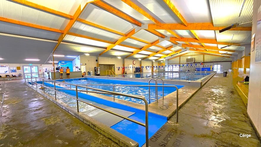 Closer view of the ramp access with steel handrails. There is a grey mat along the ramp access with small circular bumps for added friction to the feet to support the guest while entering the pool. The pool has deep blue walls, and within the cemented space surrounding the pool is the word “Caution: Slippery when wet”. A black waste bin is visible in the far-right area of the pathway. Strings of triangular blue flags can be seen hanging above the large pool.