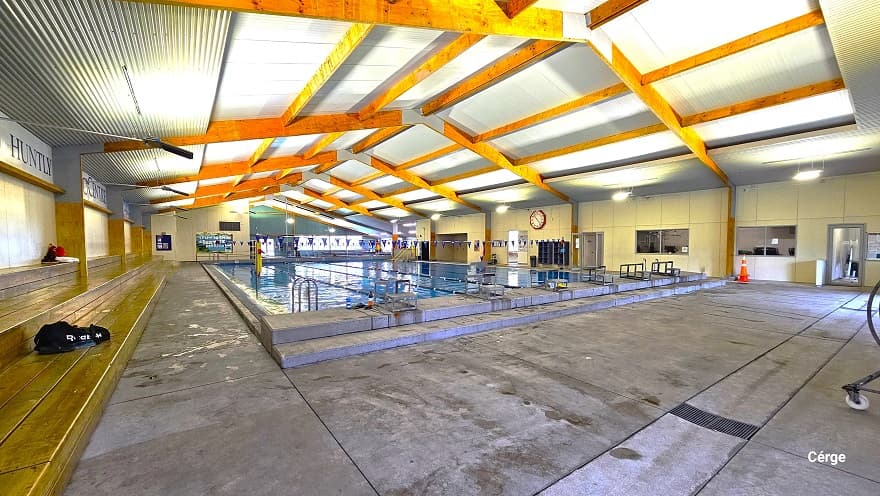 View of the large pool near the long row of low-set brown benches. On the upper wall of the back of these benches is a large font of Huntly Aquatic Centre. Opposite this wall is the wall with a large red circular clock with two open doors for bathrooms and changing rooms. Starting blocks are lined at the elevated edge.