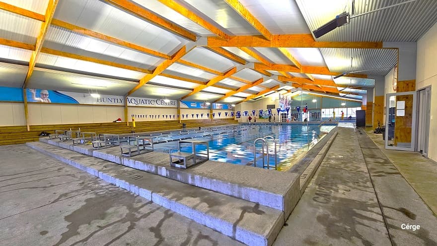 A closer look at the elevated edge at the starting point of the large pool. Grey diving blocks are placed within each pool lane. The white ceiling is corrugated designed with large wooden beams. Strings of triangular blue flags can be seen hanging above the large pool. On the right is an open glass door with small papers attached to it.
