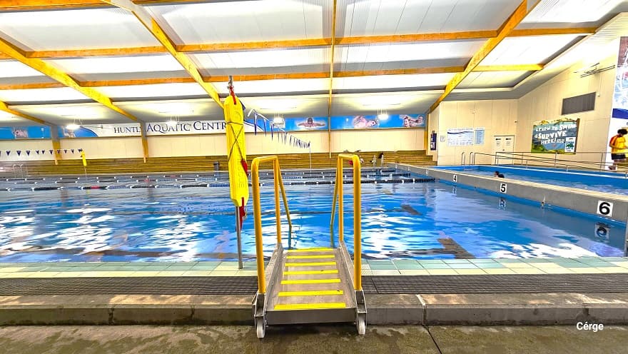 One of the access pool ladders with yellow hand railings on the side of the large pool at Huntly Aquatic Centre. On the pool wall, a few centimeters above the pool water, are numbers from 1-6 assigned for each pool lane. The wall further to the back, on the right side, is mostly covered by informative graphics. The name of the centre is clearly visible on the upper part of the wall directly opposite.