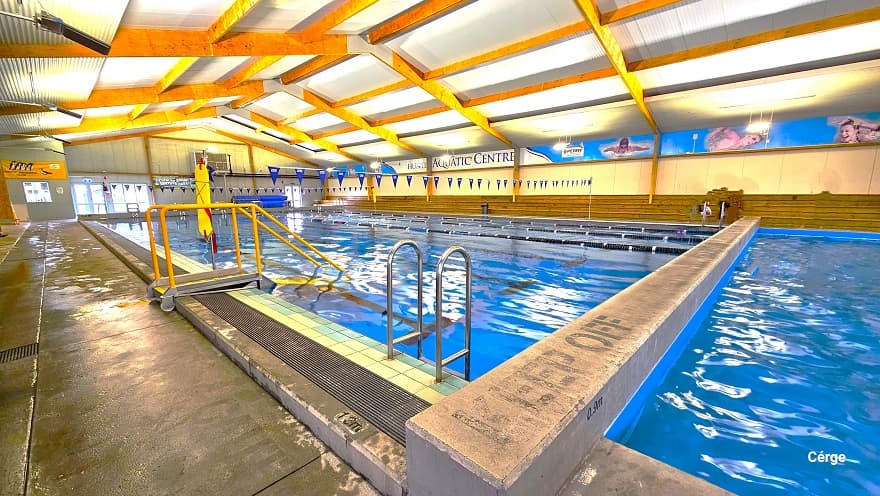 A pool wall divides the large pool and the small pools. On the top surface of this wall is a “Keep Off” phrase in black. Aside from the steel pool ladders, there is also another pool ladder but with small steel tubes of yellow handrails. Strings of triangular blue flags can be seen hanging above the large pool. Long rows of low-brown benches are available on the opposite side of this pool.