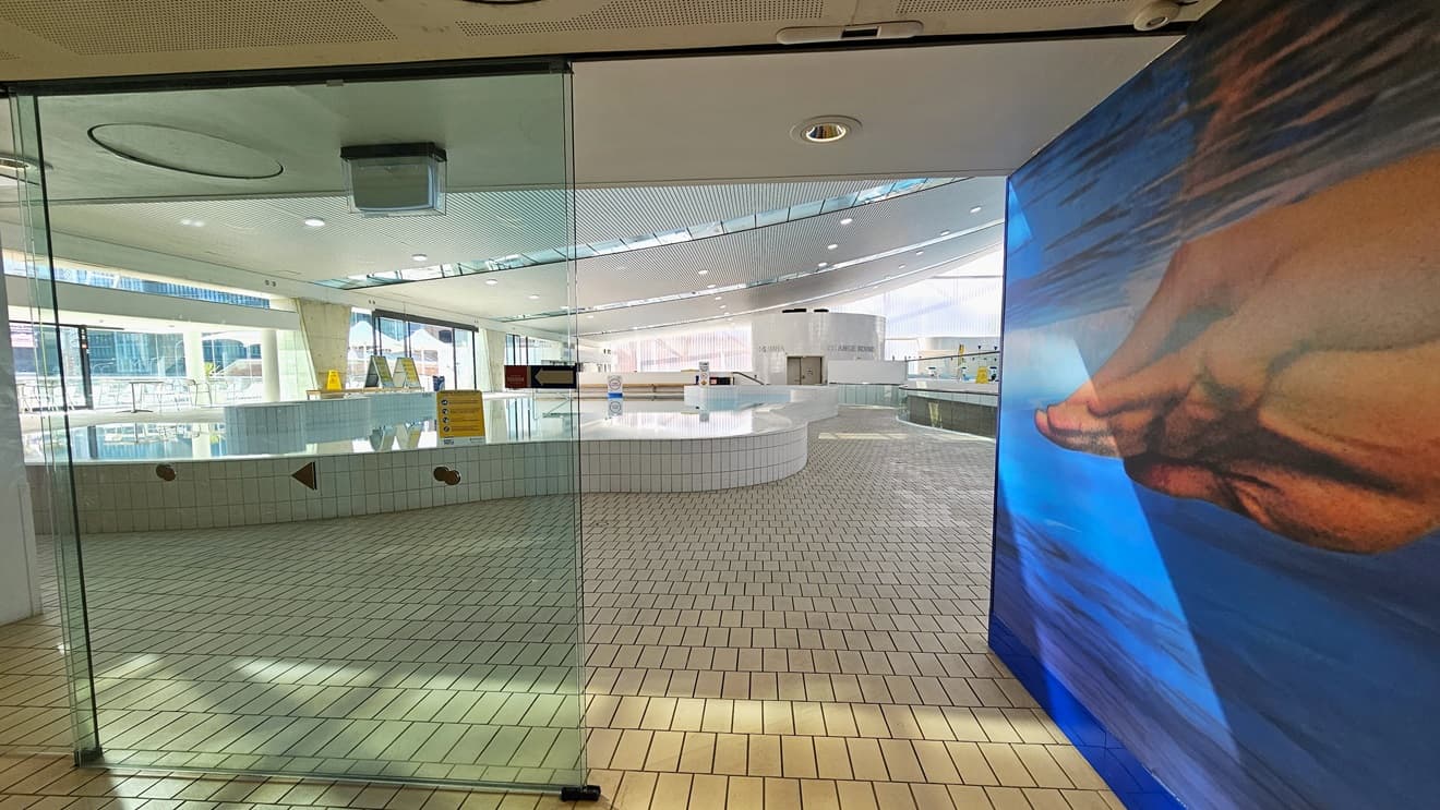 Access to the aquatic center through an automatic glass sliding door. The right wall features a life-sized painting of Ian Thorpe swimming underwater. Inside, the leisure pool with stairs and a handrail is directly ahead. The floor is tiled with small bricks, and the ceiling displays a white porous design with circular LED lights.