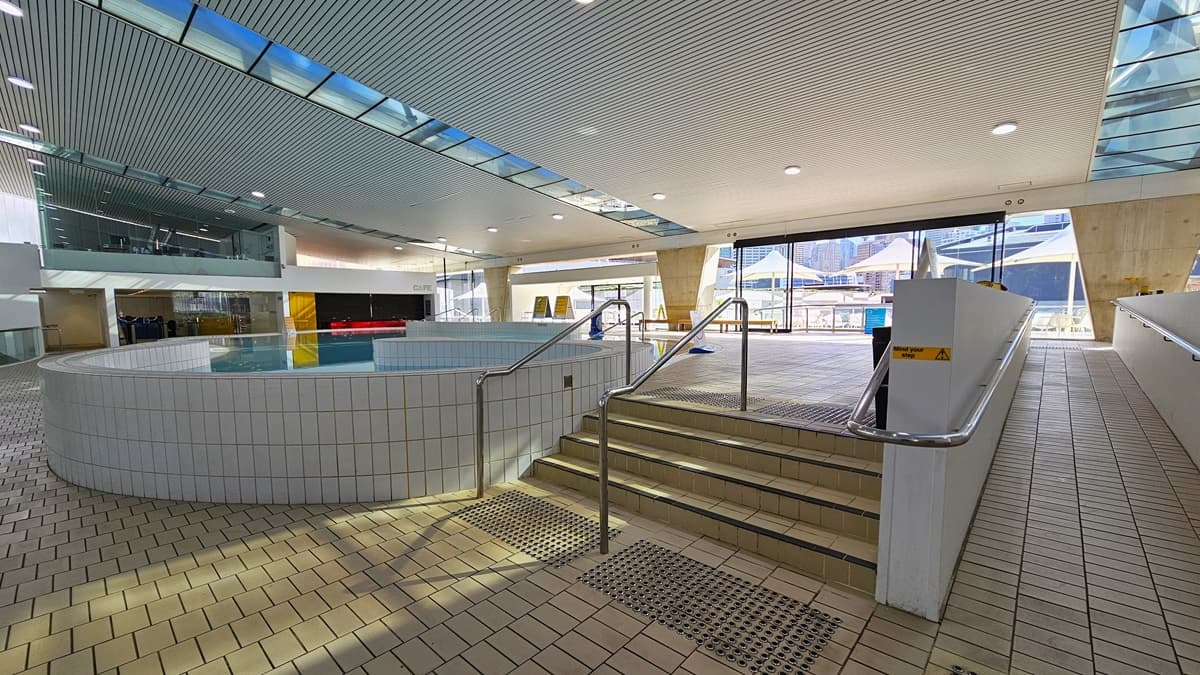 The Sauna Pool features steps with handrails and a ramp with handrails, along with truncated domes on the floor. Adjacent to it is the Program Pool, also equipped with steps and ramp entry, with visible safety signage. To the left, a glass wall provides a view of other facilities. The ground floor is tiled with small bricks, and the ceiling displays a white porous design with circular LED lights, incorporating clear glass reflecting the pool below. Beyond lie the sauna and Program Pool.