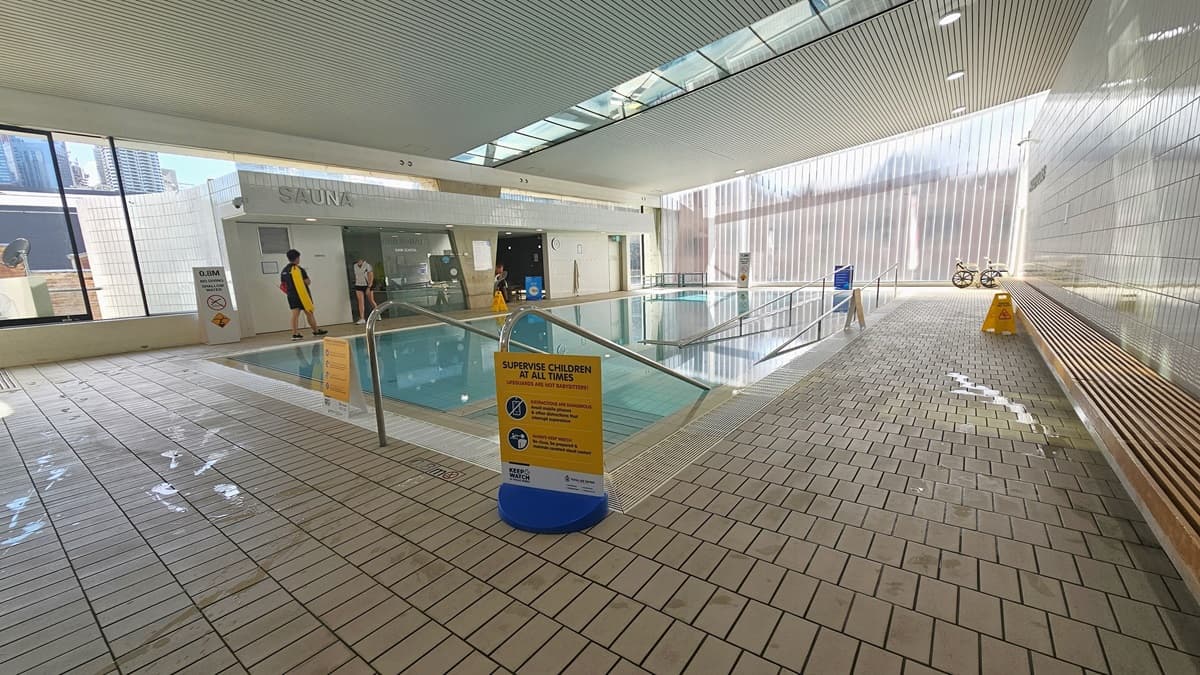 The Program Pool is furnished with steps and a ramp with handrails, accompanied by a ramp with handrails. Freestanding banners are affixed to the pool deck. A long wooden bench is situated against the right wall, and aquatic wheelchairs are positioned in the corner. The ground floor is tiled with small bricks, and the ceiling exhibits a white porous design with circular LED lights, featuring clear glass glass reflecting the pool below. Beyond lies the sauna and Program Pool.