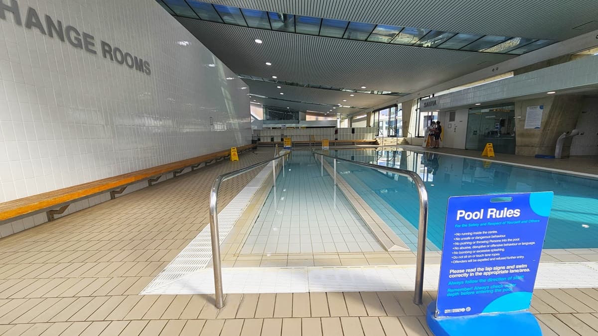 The Program Pool is equipped with ramp entry and handrails. Freestanding banners displaying Pool Rules and safety warnings are affixed to the pool deck. A long wooden bench is positioned against the left wall. The ground floor is tiled with small bricks, and the ceiling features a white porous design with circular LED lights, reflecting the pool below. To the left is a white brick wall with a sign indicating "Change rooms."