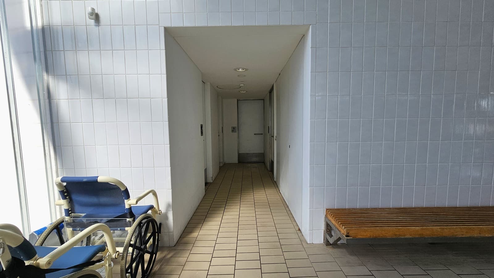 Entrance to the accessible bathrooms. Two aquatic wheelchairs are positioned on the left, and a long wooden bench is on the right. The wall is made of small bricks. The corridor leading to the bathrooms has cream floors, white walls, and white ceilings with circular LED lights.