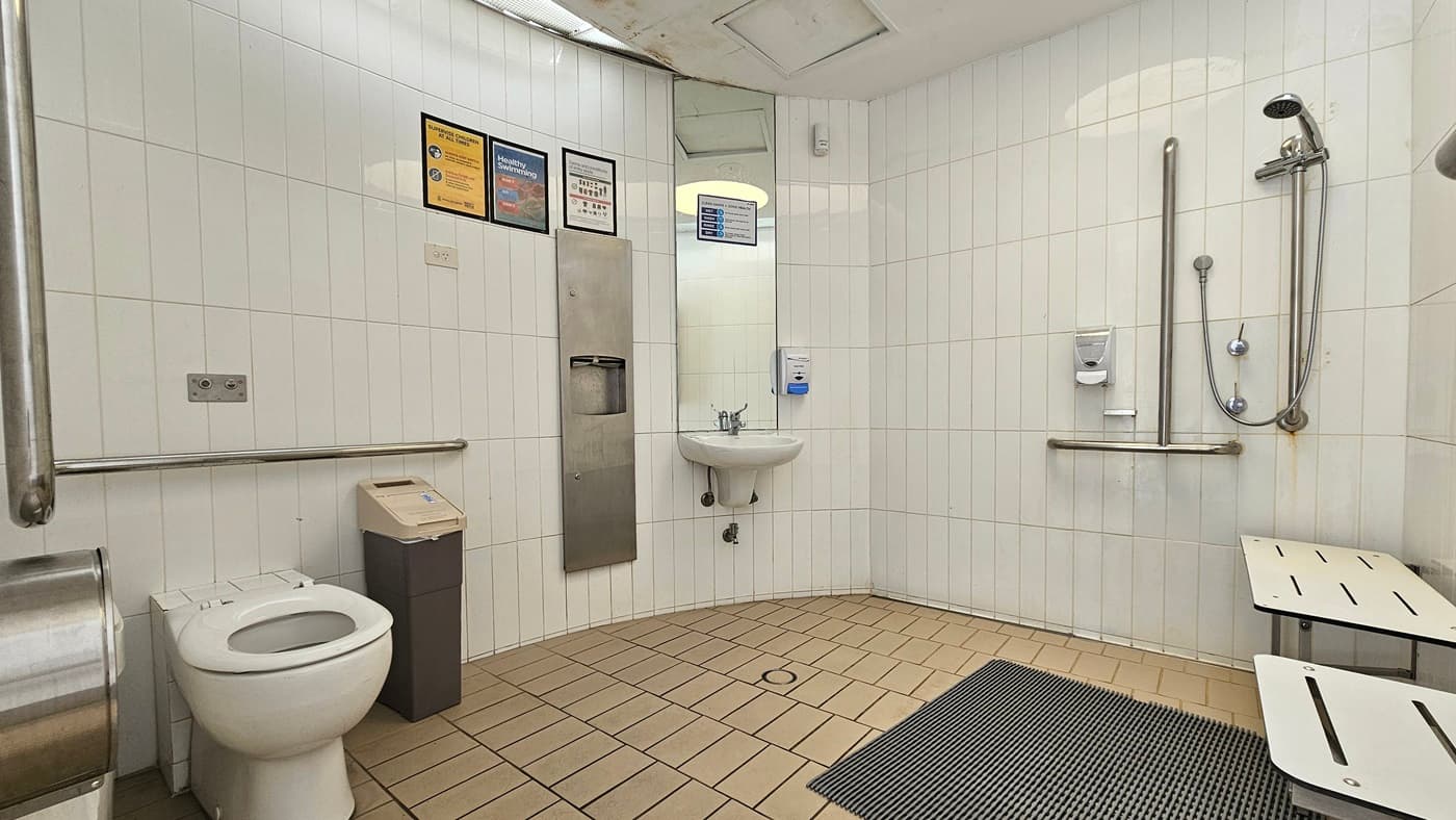Within the accessible bathroom, a toilet bowl with hand grabs is situated on the left, accompanied by a tissue dispenser and a rubbish bin. Adjacent to it on the left wall are a wash basin, tissue dispenser, mirrors, and soap dispenser. On the right, there is a shower with handrails and a foldable bench is placed on the right wall. Banners are affixed to the upper left wall. The walls are adorned with white tiles, and the floor is covered in brown tiles with a non-slippery mat.