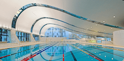 The white ceiling displays a wave-designed porous pattern with glass structures following the ceiling contours and is equipped with LED lights. The glass structures reflect the pool below, adorned with colourful floating ropes. The left side has glass windows providing a view of the outside.