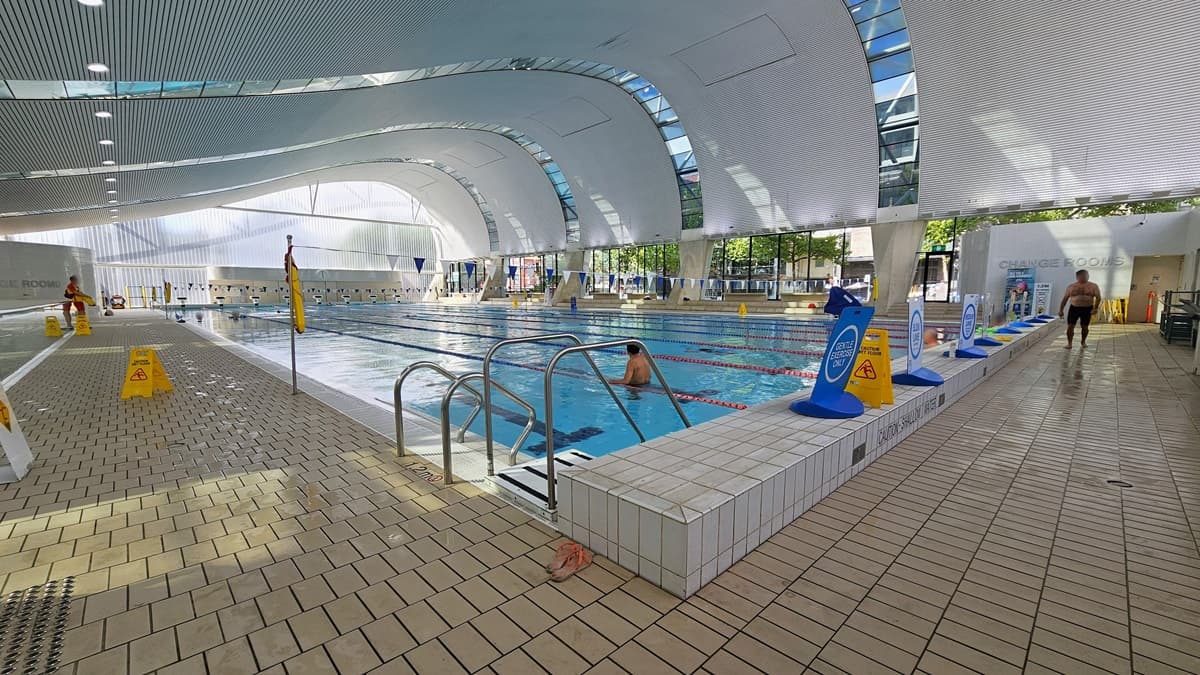 The Lap pool features steps with handrails, floating ropes, and blue and white buntings running across the pool. Freestanding banners line the raised pool deck. The change room is on the right, and safety warning signs and truncated domes are on the left pool deck. Some guests are using the pool. The ground floor is tiled with small bricks, and the wave-shaped ceiling displays a white porous design with circular LED lights, reflecting the pool below.