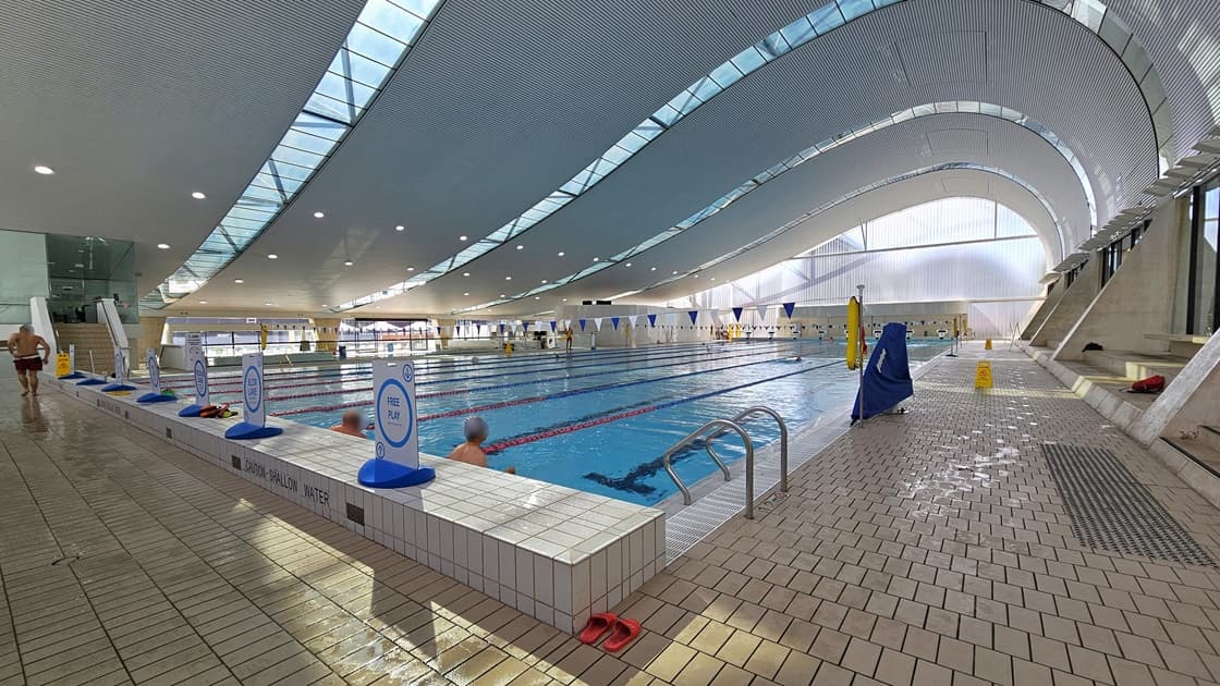 The Lap pool has steps with handrails, equipped with truncated domes. Floating ropes and blue and white buntings run across the pool. Freestanding banners line the raised pool deck on the left. Some guests are using the pool. The floor is tiled with small bricks, and the wave-shaped ceiling features a white porous design with circular LED lights, reflecting the pool below.