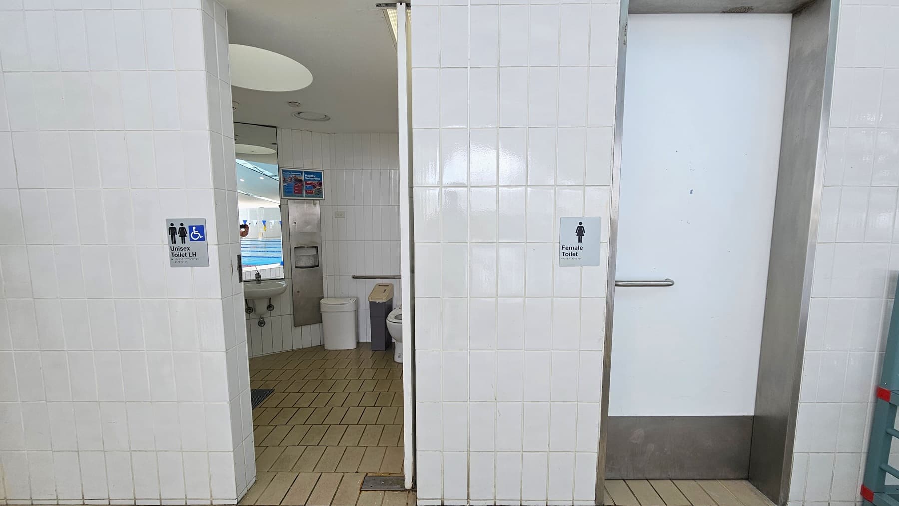 The left door leads to an all-gender and accessible toilet. Inside, the toilet bowl is positioned against the wall, accompanied by rubbish bins, mirrors, a washbasin, and a tissue dispenser. The floor is covered in brown tiles, the walls are adorned with white tiles, and the ceiling features circular LED lights. The left door designates a female toilet.