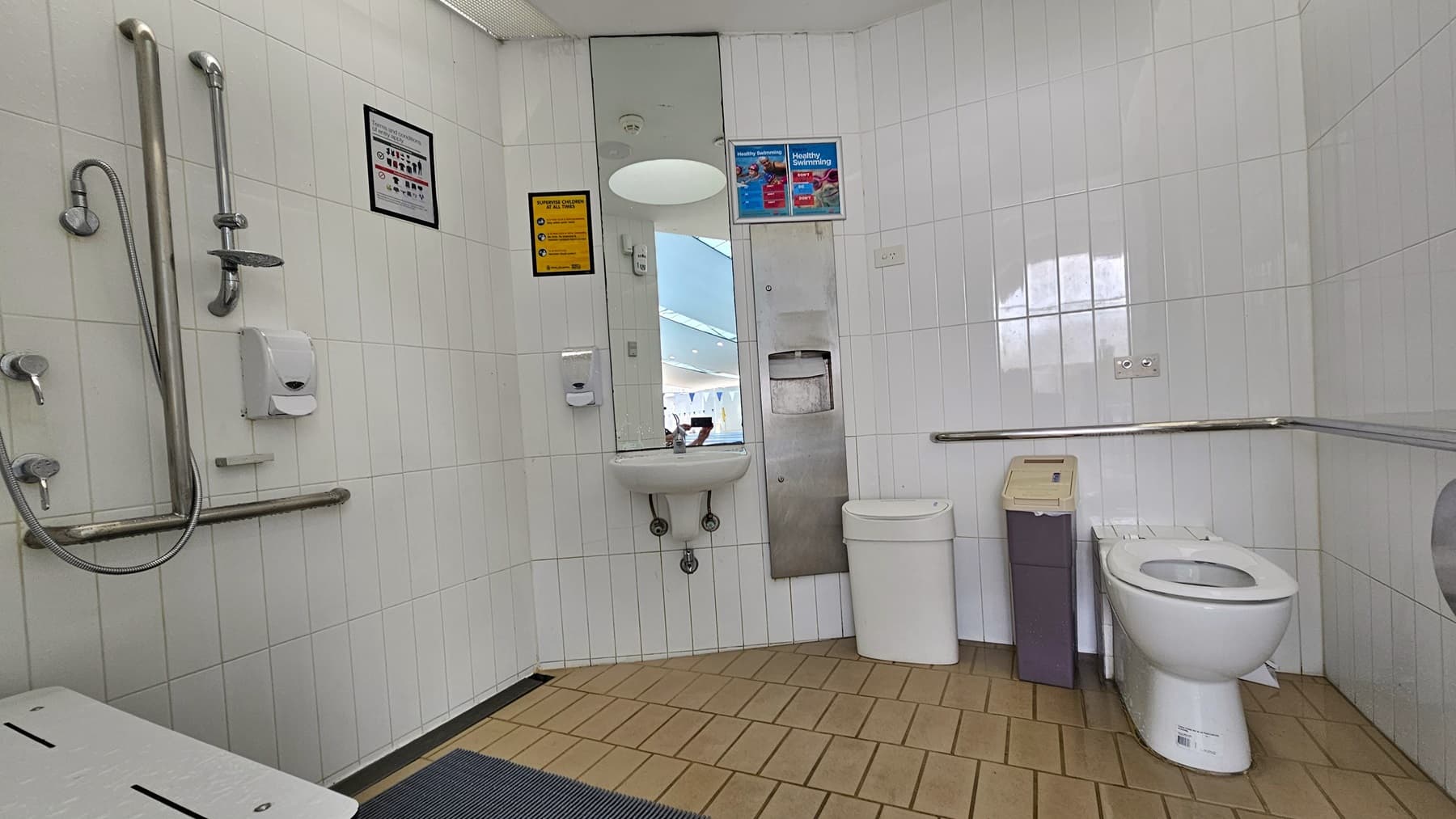 Within the accessible bathroom, the toilet bowl is against the right wall with accompanying rubbish bins. Mirrors, a washbasin, and a tissue dispenser are mounted on the left wall, along with showers featuring handgrabs. Adjacent to these fixtures is a foldable bench. The floor is covered in brown tiles with a non-slippery mat, and the walls are adorned with white tiles. The ceiling is equipped with circular LED lights. The left door designates a female toilet.