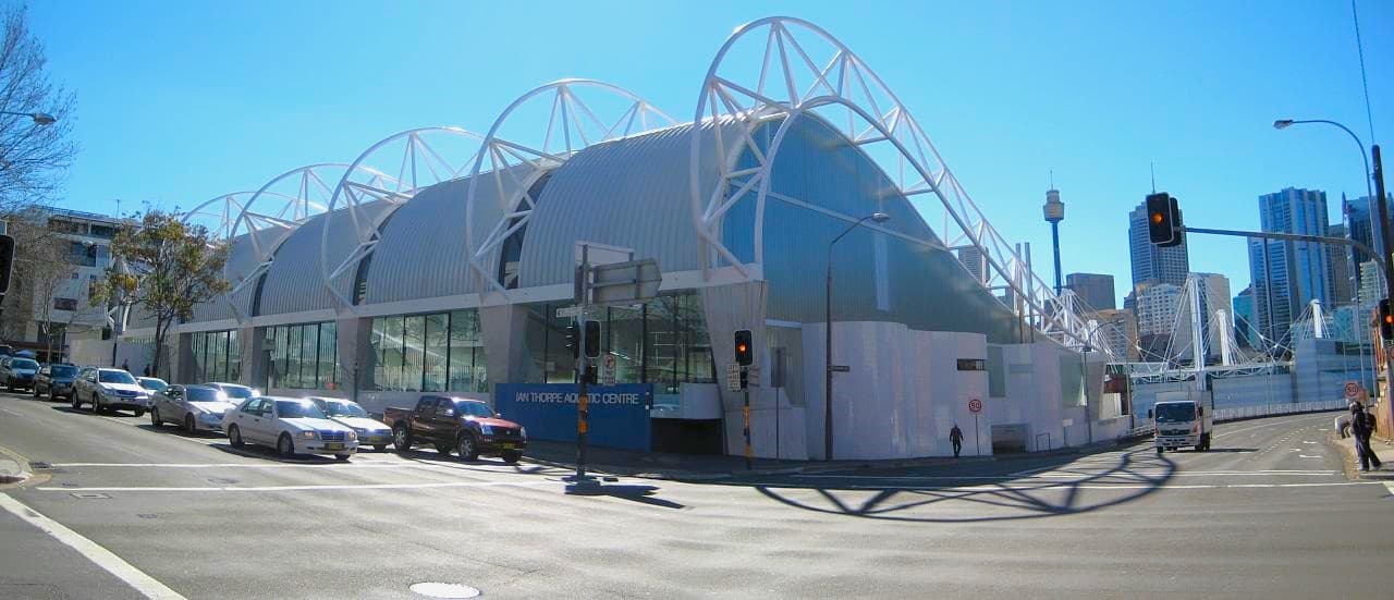 At a road intersection stands a modern commercial building with a unique blue corrugated wave roofing design. White metal structures complement the roofing contours. A blue wall displaying the facility sign is on the left. To the right, a stop light is visible, with cars stopped on the left side. Further back, tall commercial buildings can be seen.