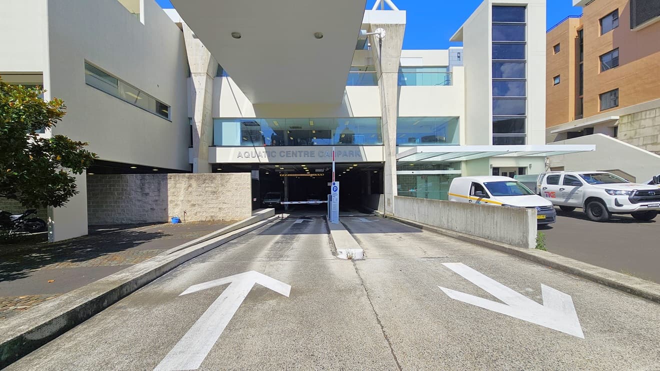 The two-way road provides access to and from the underground car park. The facility name is displayed before the entrance, separated by a traffic median. Limited parking is available on the right, with two cars currently parked. A brown facility building is situated on the right, and a tree is visible in the concrete area on the left.