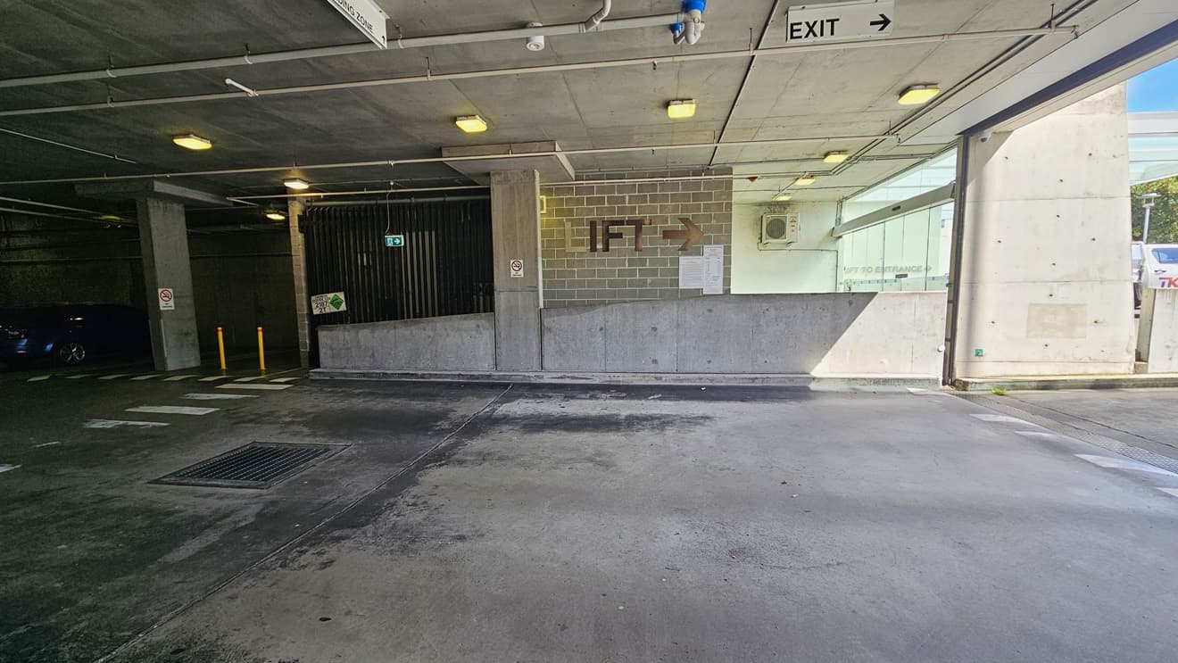 The underground parking includes a pedestrian lane on the left, marked by yellow bollards. The middle brick wall has a sign indicating the lift direction. The low ceiling features directional signs, LED lights, and exposed pipes. Safety signs are affixed to the wall, and a visible water drainage system is present.