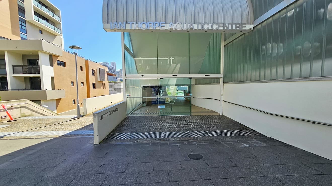 The facility entrance is through an automatic glass sliding door, with truncated domes on the floor. The right wall features a glass structure, and the facility name is written on top of the wave-shaped roofing. To the left is a multi-story cream building, with a direction leading to the car park visible on the left.