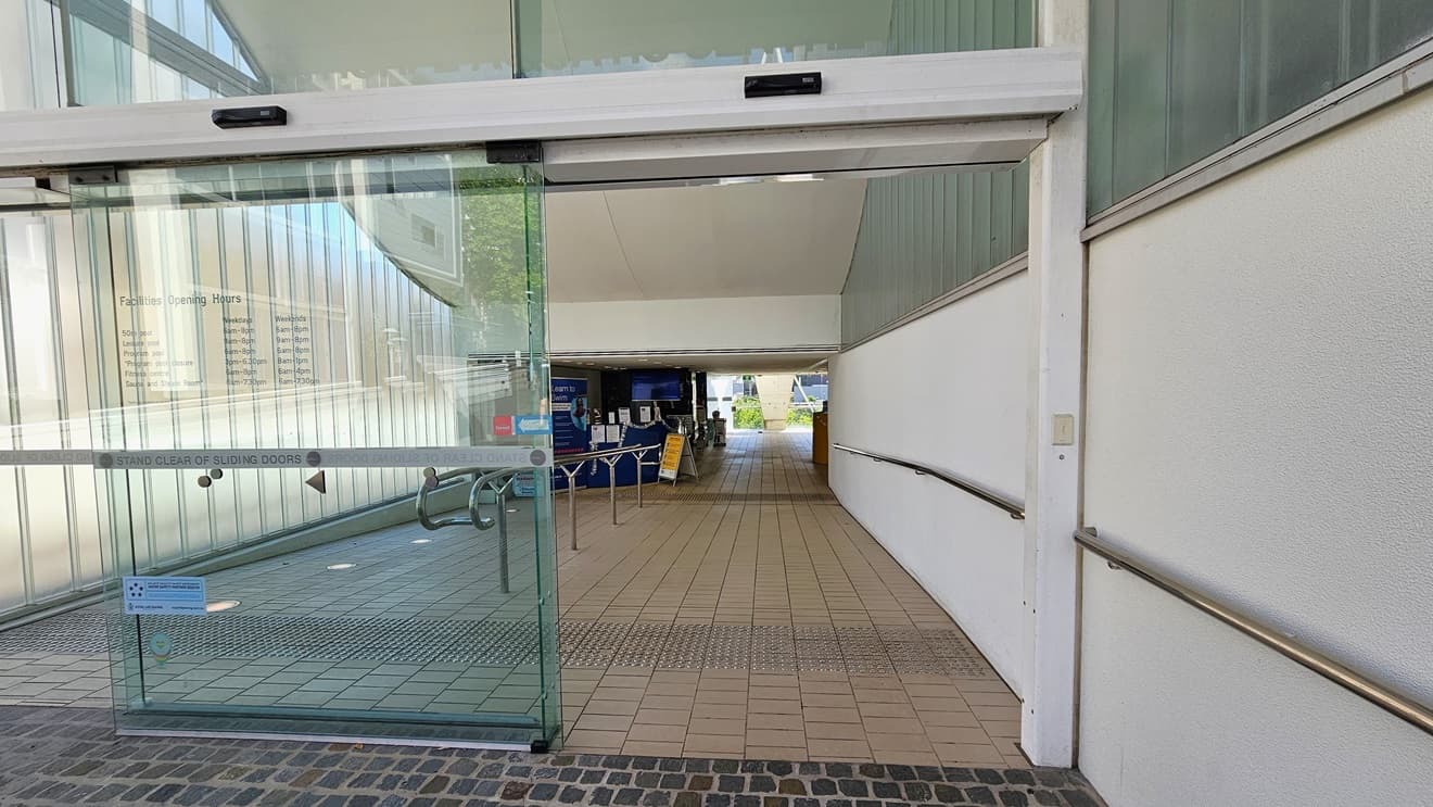 The automatic glass sliding door displays the facility sticker. Truncated domes are installed on the floor. The lobby path has a gentle descent, with handrails positioned in the middle and on the right. Moving ahead, the reception is visible. The floor is covered with grey tiles.